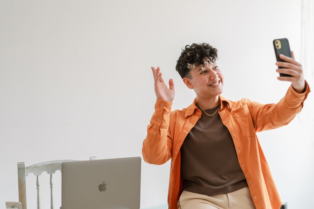 Marketing by Rocio founder and SEO and AI specialist Rocio Sanchez sitting on desk while waving at smartphone screen with laptop next to them in 2024.