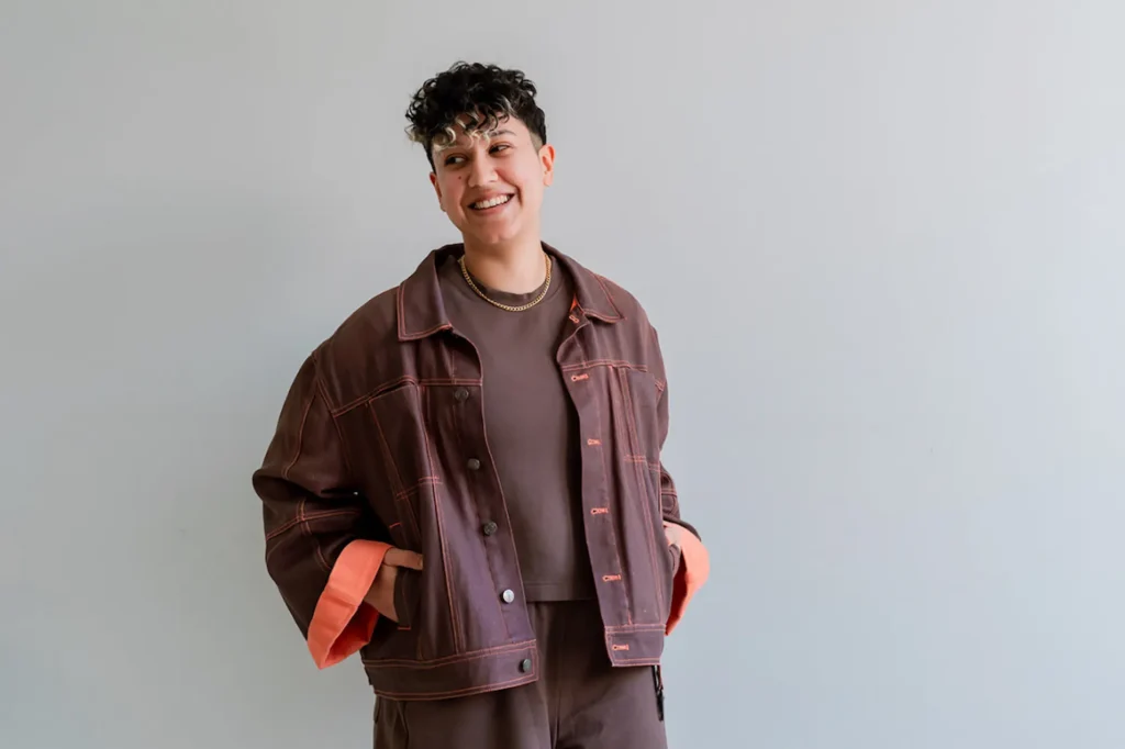 Marketing by Rocio founder Rocio Sanchez in a brown oversized denim jacket and gold chain necklace standing against a light gray background