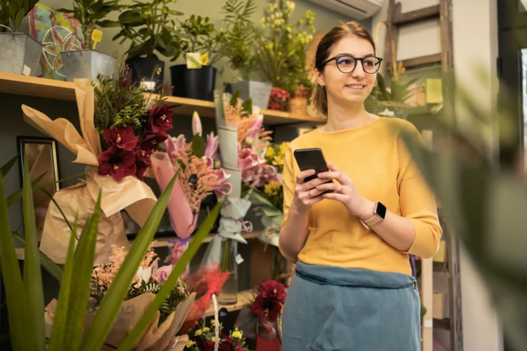Small business owner standing in a flower shop holding a smartphone and smiling.