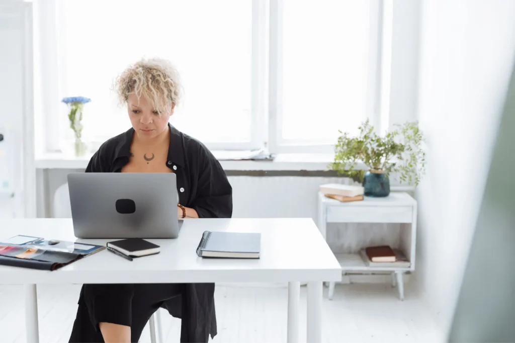 A person with short, curly blonde hair sitting at a clean white desk, focused on working on a laptop in a bright, minimalist room.