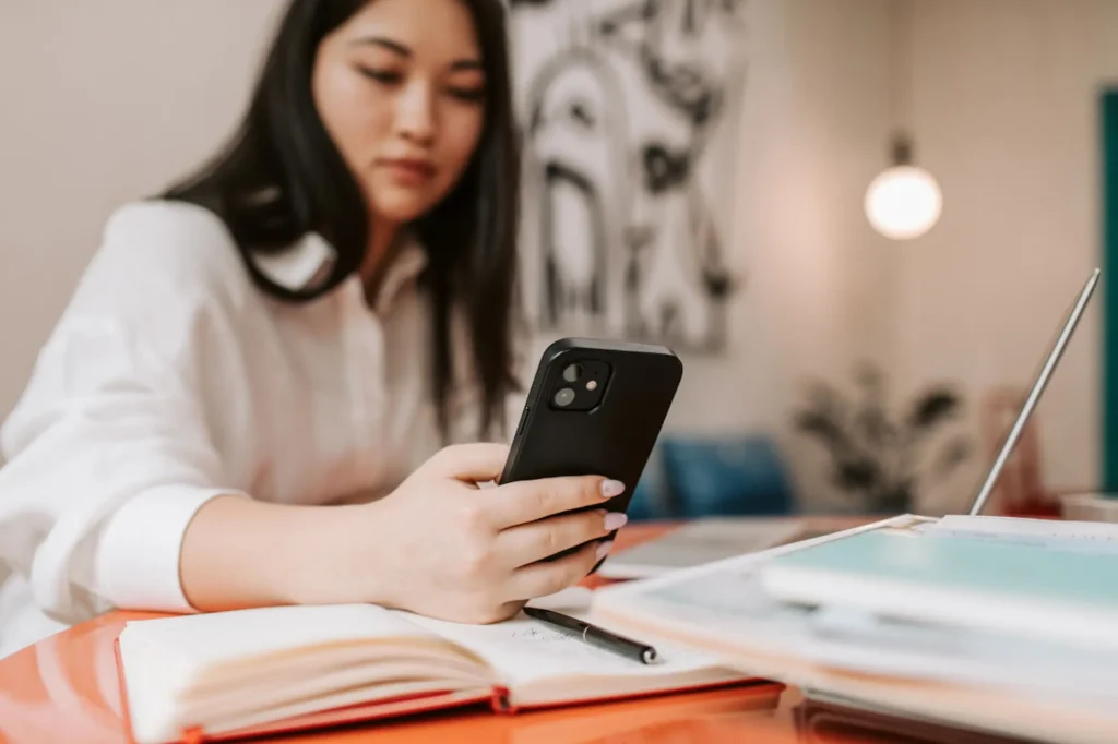 Business owner checking social media on a smartphone while working at a desk with an open notebook and laptop