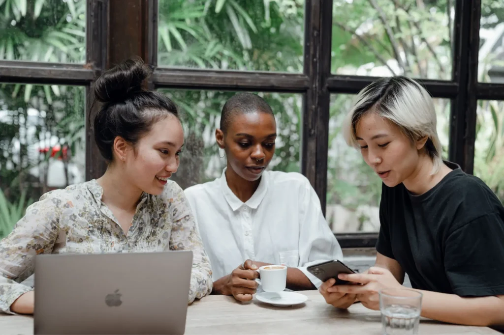 Three business owners sitting together at a wooden table near a large window with green plants outside. One looks at a laptop, while the other two look down at a smartphone