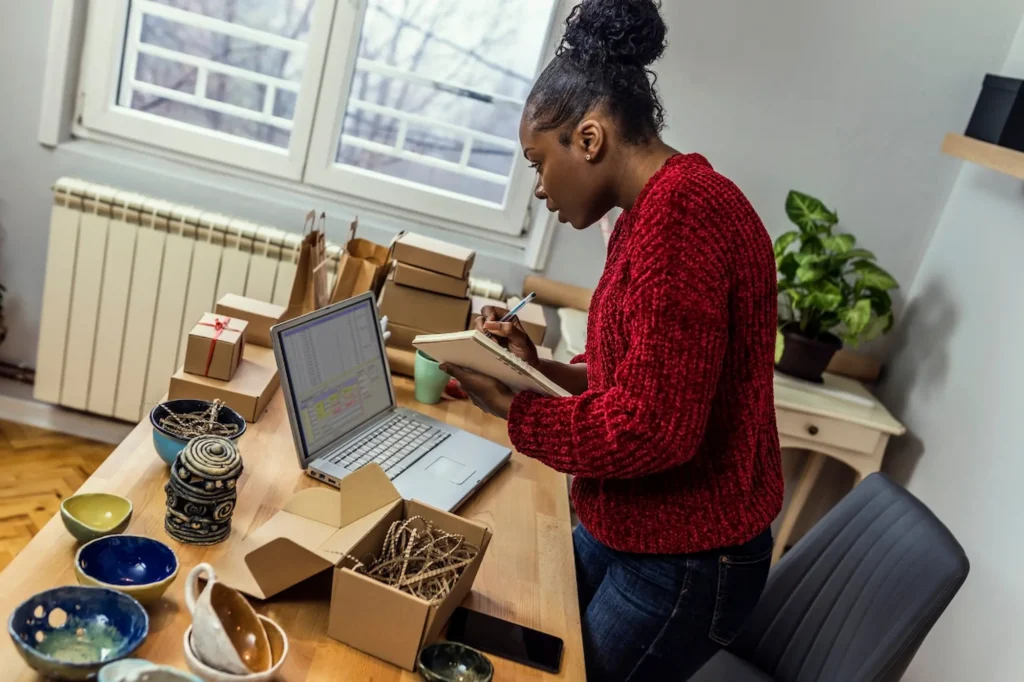 Black woman small business owner taking notes beside her laptop and handmade ceramic products while planning her blog content strategy