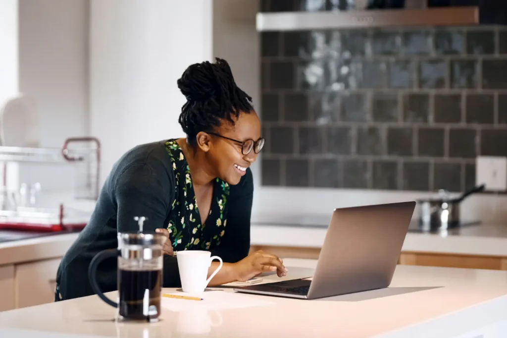 Black businesswoman smiling while reading a blog post on her laptop over morning coffee in her kitchen