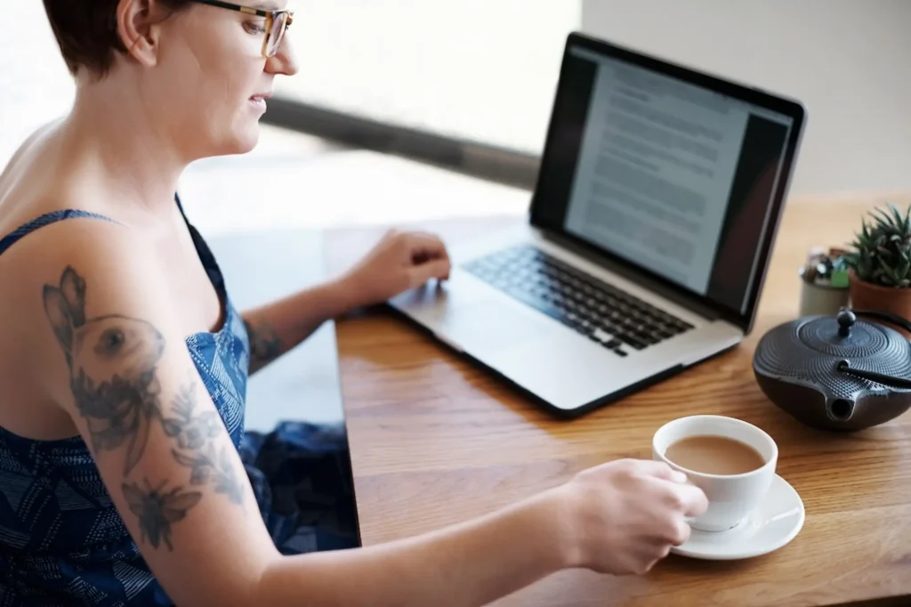 Small business owner with tattoos writing a blog post on her laptop while enjoying tea at a café workspace