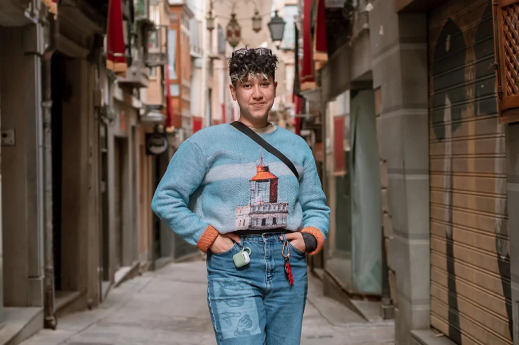 Marketing by Rocio founder Rocio Sanchez standing confidently in a narrow city alley wearing a graphic knit sweater, representing authentic queer representation in advertising campaigns for 2026.