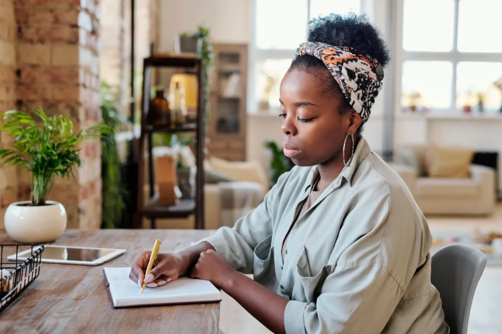 Black woman small business owner writing blog ideas in a notebook at her home office desk surrounded by plants and natural light