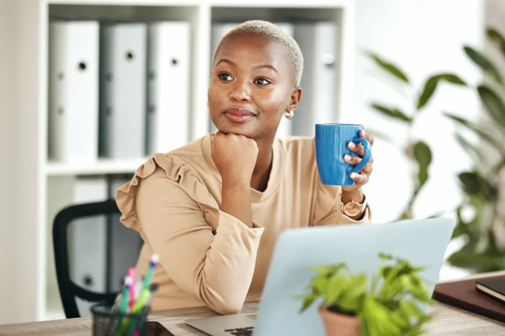 Black business owner sitting at her desk with a laptop, holding a coffee mug and looking thoughtfully to the side