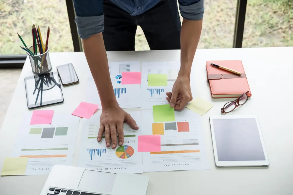 Person standing over a desk covered in printed charts, graphs, and sticky notes while reviewing marketing data