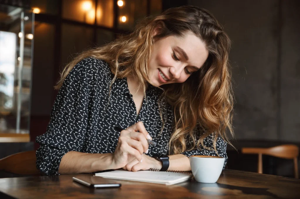 Woman blogger smiling while writing blog post ideas in a notebook over coffee at a café workspace