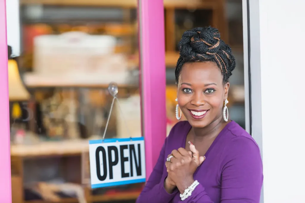 Smiling Black business owner in a purple top standing in her shop doorway beside an “Open” sign