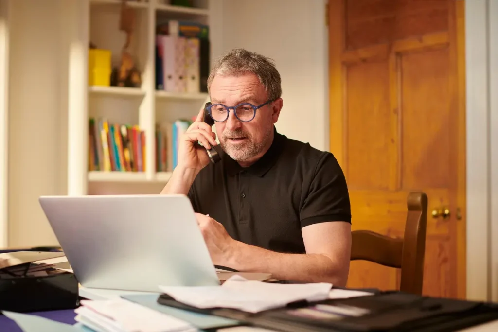 Small business owner on a phone call while reviewing his website on a laptop at a home office desk