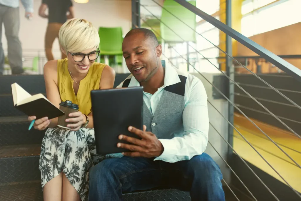 Two colleagues sitting on a staircase, smiling and looking at a tablet together