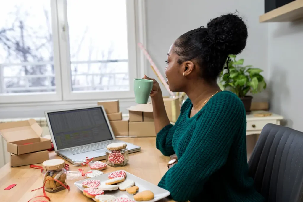 Small business owner drinking coffee at her desk beside a laptop, packaging boxes, and decorated cookies